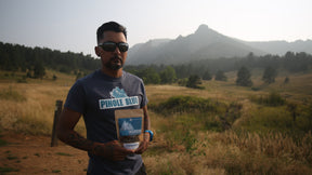Athletic man wearing sunglasses holding a package of endurobites while standing in front of a mountain.